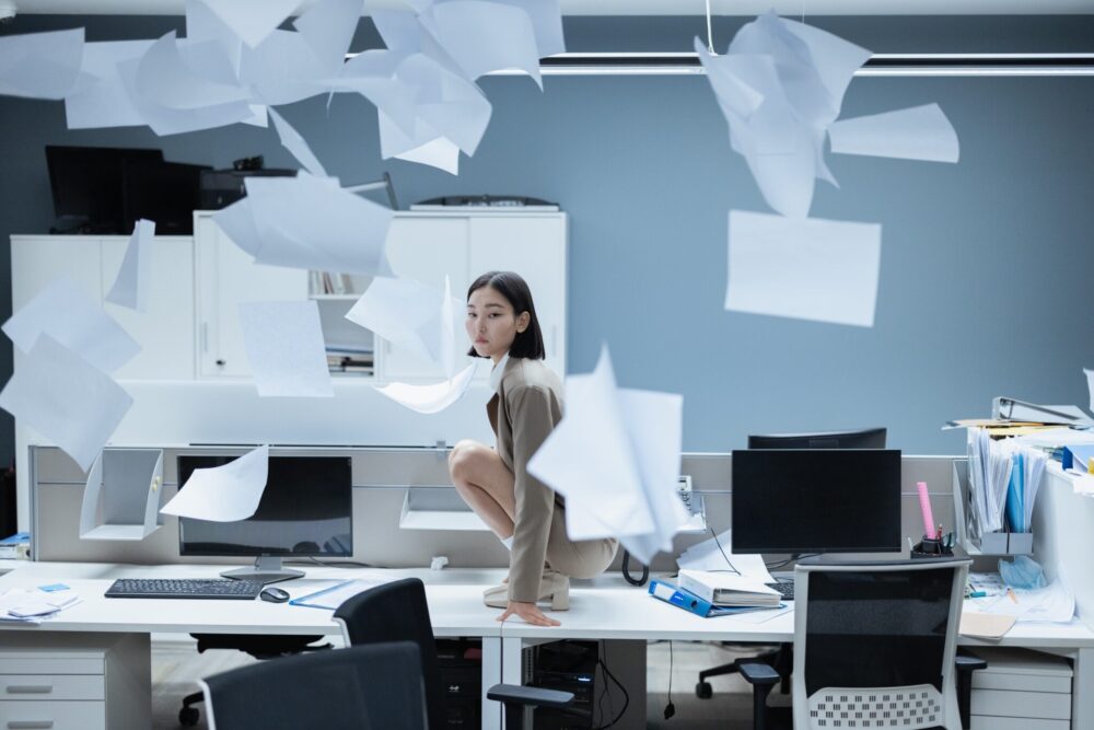 Person crouching on office desk, papers flying everywhere, chaotic scene.