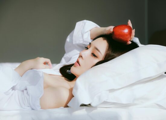 Person lying on bed, holding a red apple, white sheets.