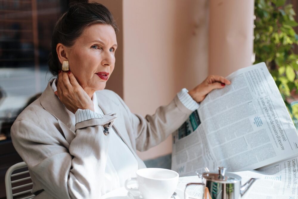 Person sitting at table, reading newspaper, drinking coffee, looking thoughtful.