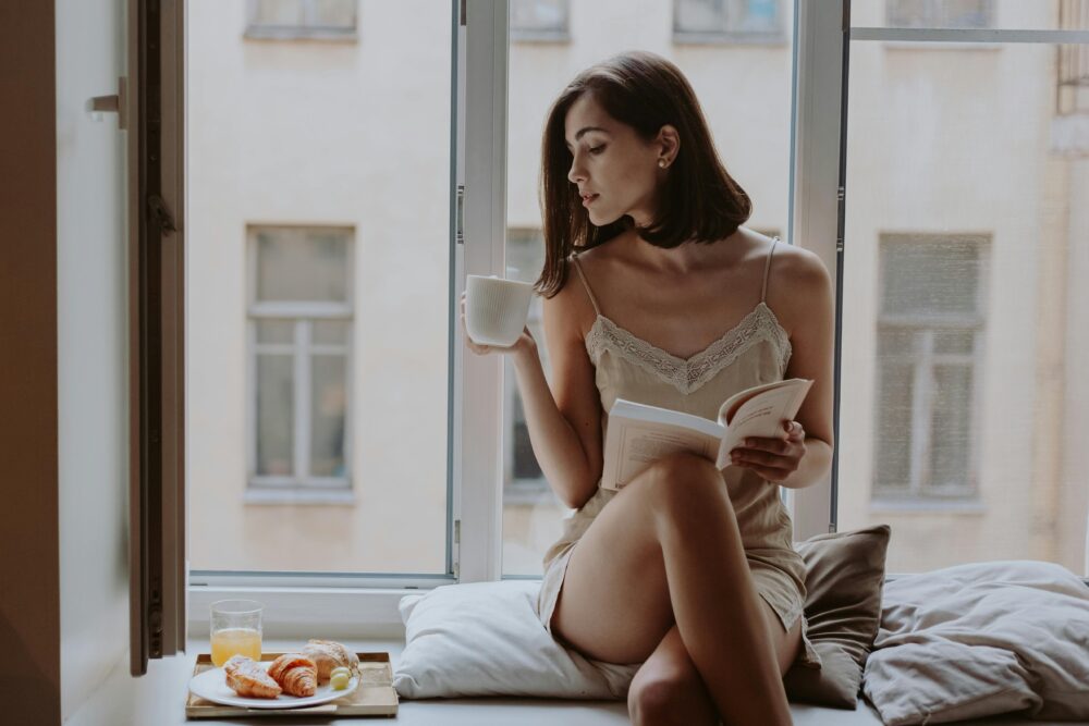 Person sitting by window, drinking coffee, reading book, breakfast nearby.