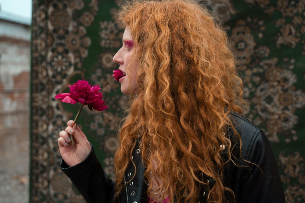 Person with red hair holds a pink flower to their mouth.