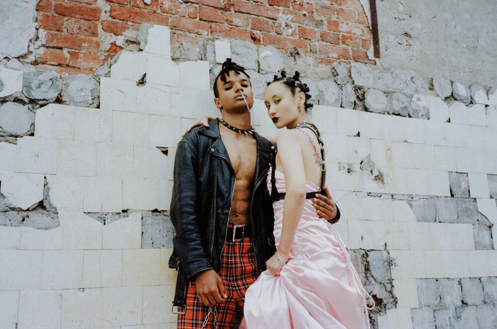 Two people in edgy fashion stand against a distressed brick wall.