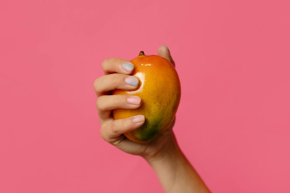 A hand holding a mango against a bright pink background.