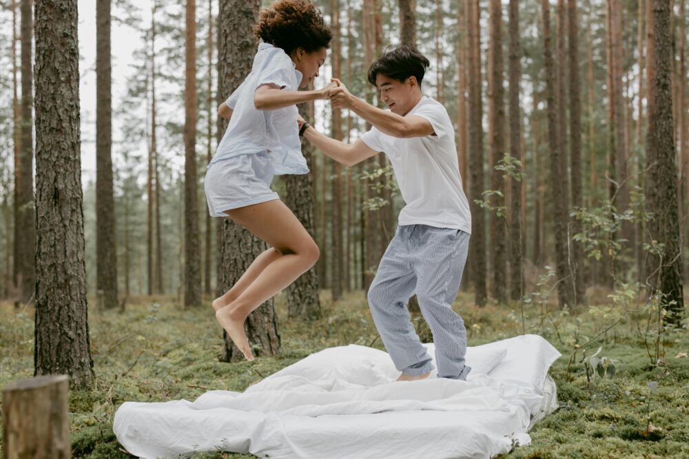 Two people jumping on a bed outdoors in a forest.