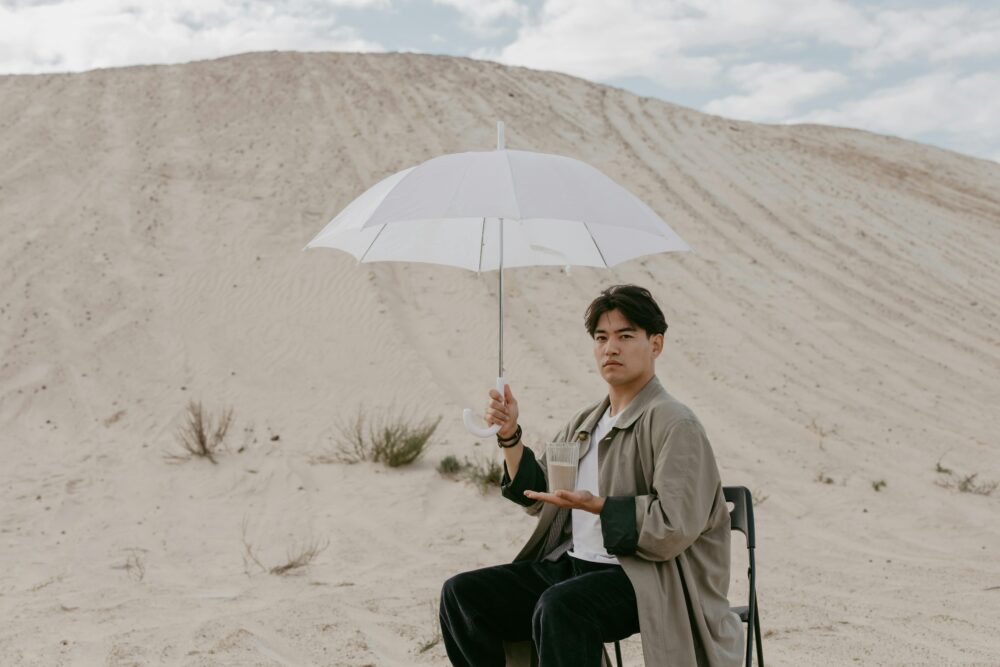 Person sitting in desert holding umbrella and glass of water.