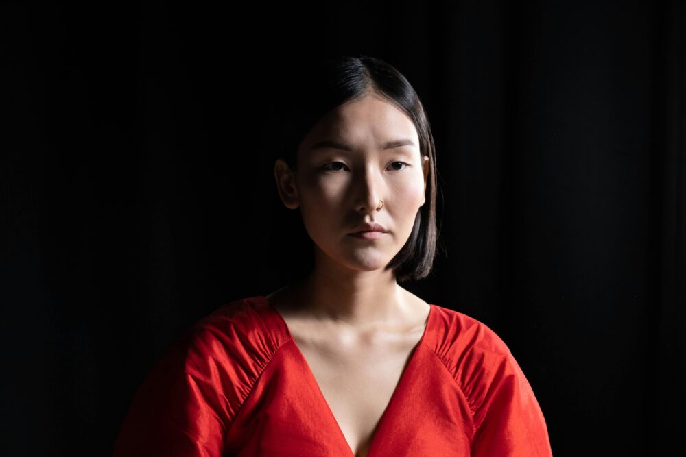 Person in red top against dark background, neutral facial expression.