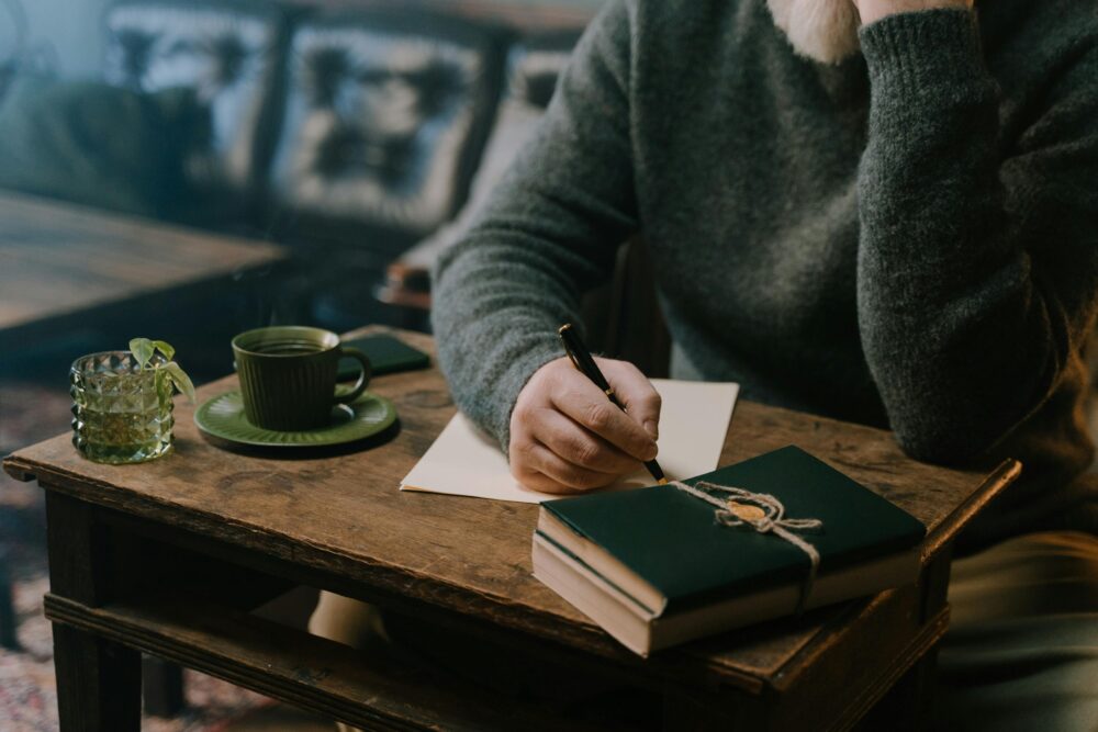 Person writing at a wooden table with drinks and books.