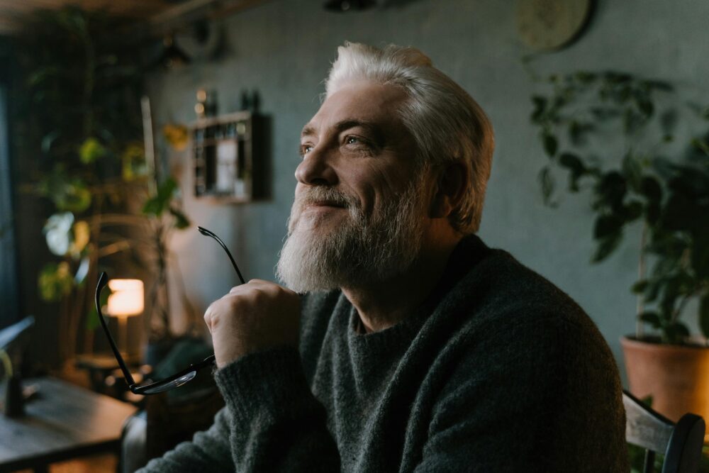 Person smiling thoughtfully indoors, holding glasses, surrounded by plants.