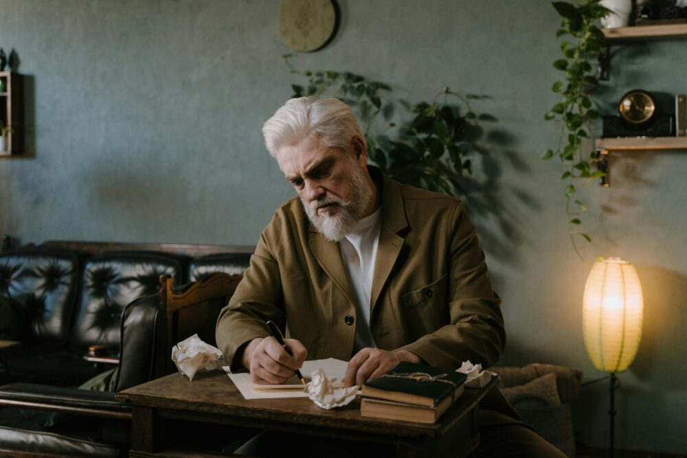 Person writing at desk, surrounded by books and crumpled paper.