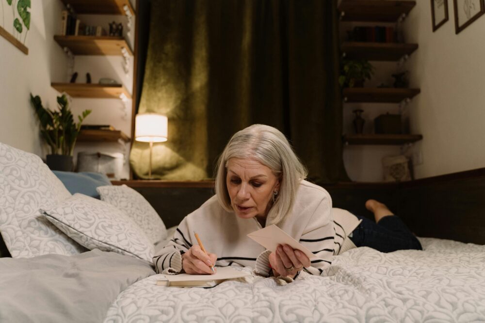 Person lying on bed, writing in notebook, holding envelope, cozy room.