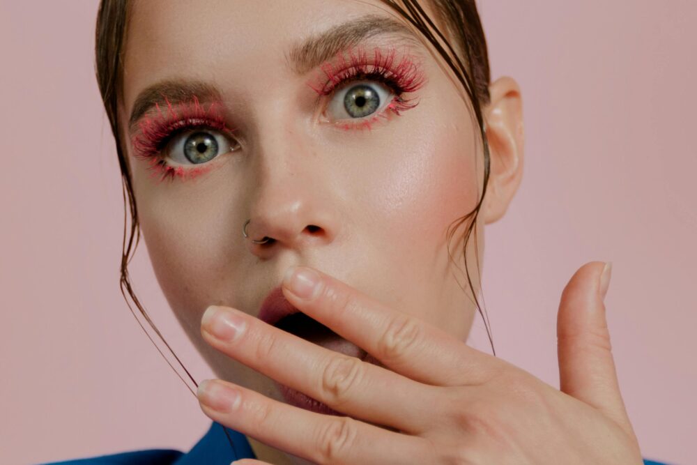 Person with surprised expression, pink makeup, hand near mouth.