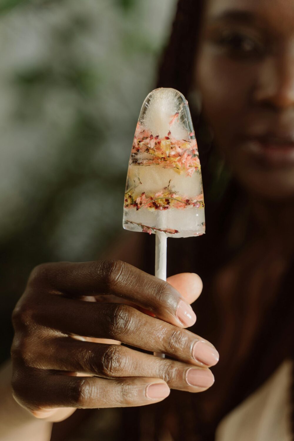 Hand holding a popsicle with flowers frozen inside, outdoors.