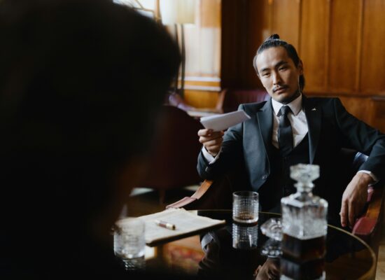 Man in suit offers paper, whiskey decanter and glass on table.
