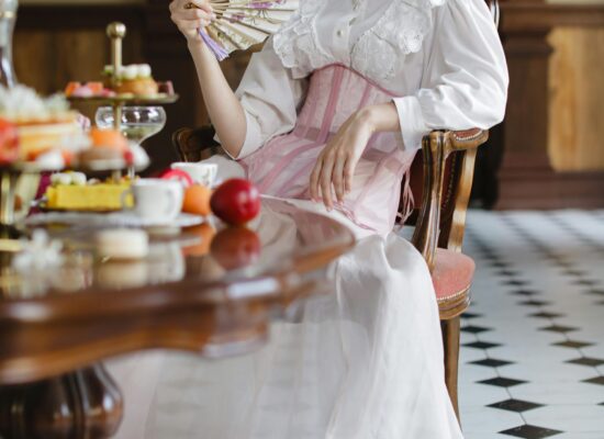 A person in vintage attire holds a fan near a table with desserts.