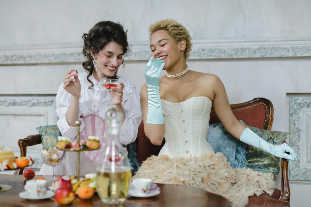 Two people in vintage attire enjoy a tea party with pastries and drinks.