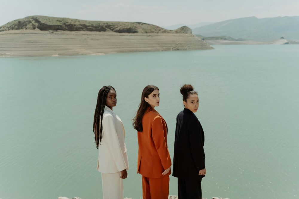 Three people in suits stand by a calm lake with hills.
