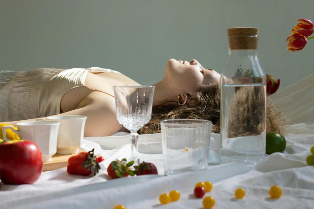 Person lying on white cloth, surrounded by fruit and glassware.