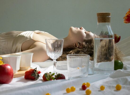 Person lying on white cloth, surrounded by fruit and glassware.