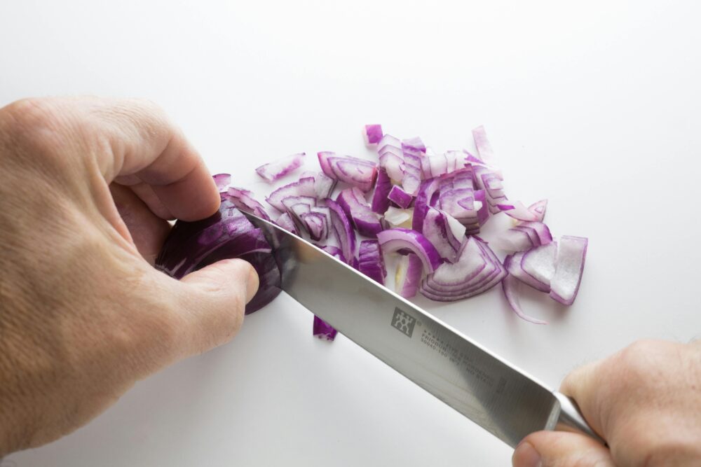 Hands chopping a red onion with a knife on white surface.