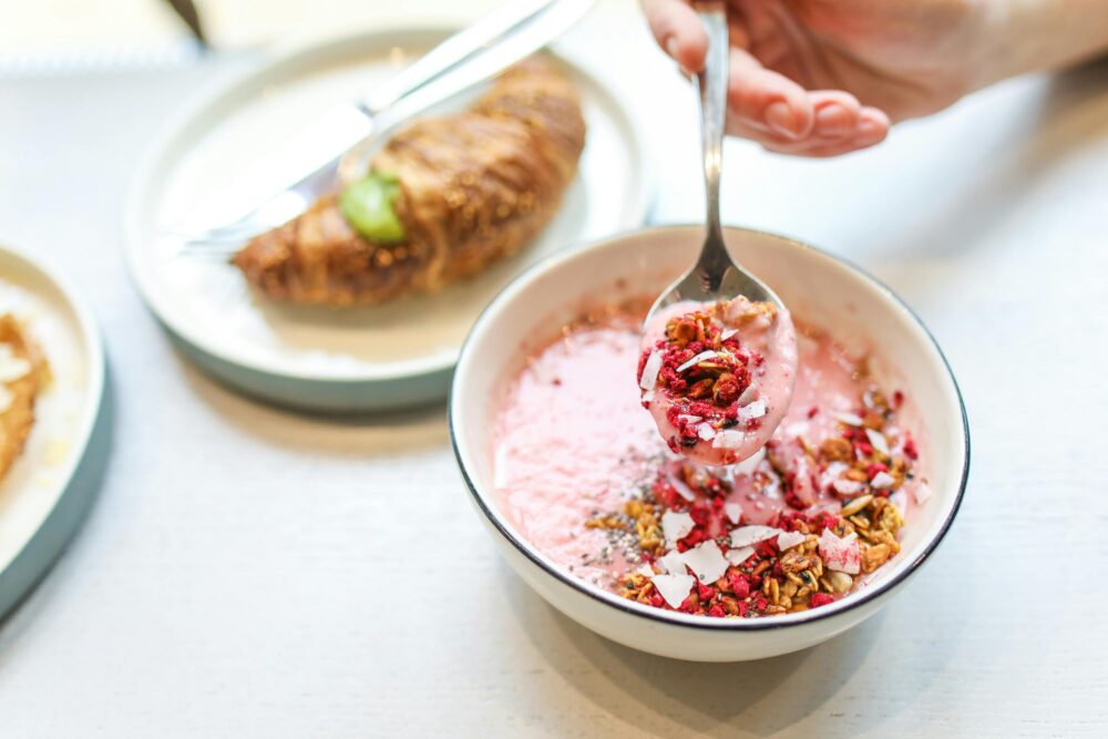 Bowl of pink yogurt with granola, croissant in background.