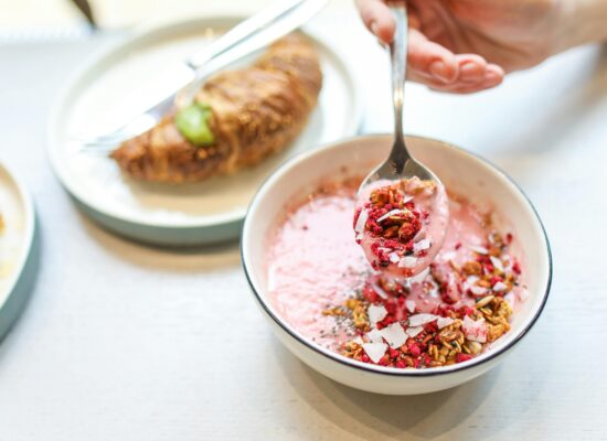 Bowl of pink yogurt with granola, croissant in background.
