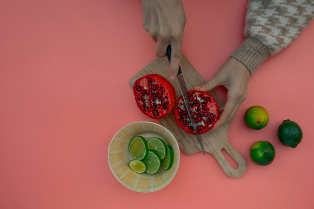 Hands cutting pomegranate, limes in bowl, pink background, wooden board.