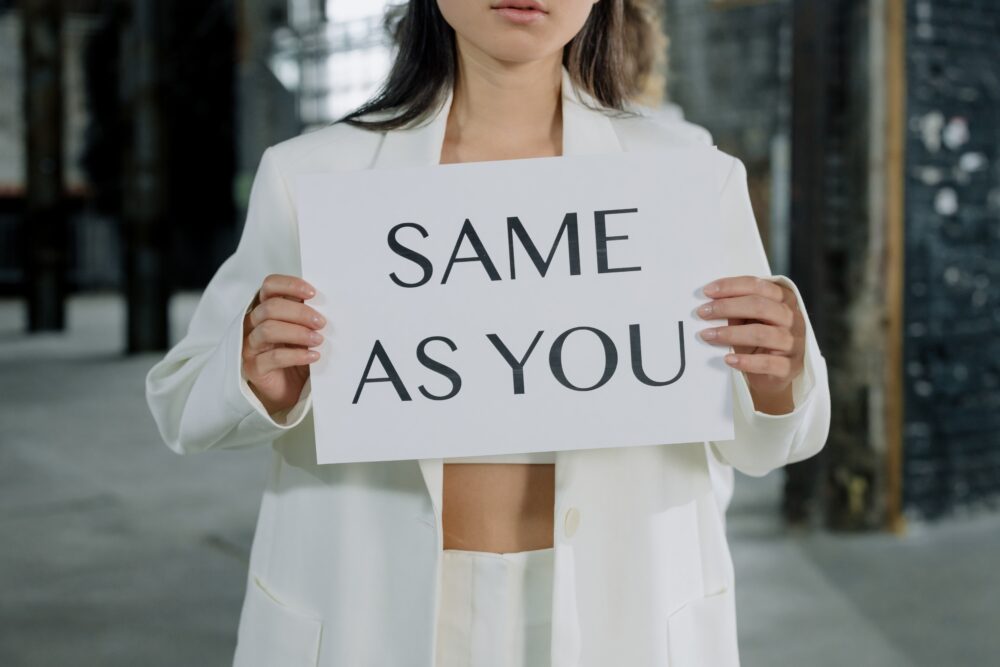 Person in white suit holding sign saying 'SAME AS YOU'.