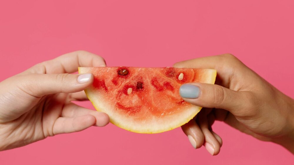 Two hands holding a watermelon slice against pink background.