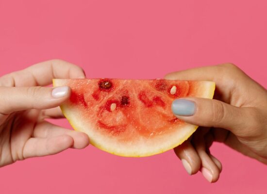 Two hands holding a watermelon slice against pink background.
